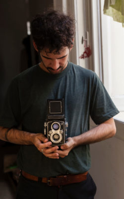 Young curly haired alternative photographer using an old medium format film camera brandless on a studio interiors with natural light. Young people using analog photography as a way to follow this art Young curly haired alternative photographer using an old medium format film camera brandless on a studio interiors with natural light. Young people using analog photography as a way to follow this art